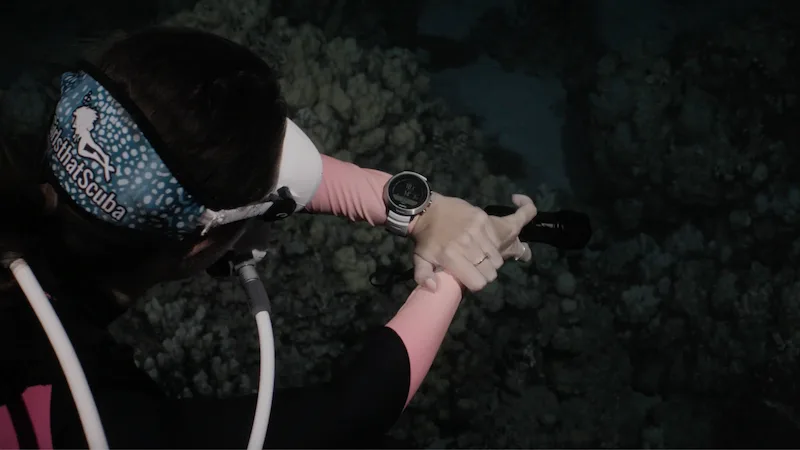 A scuba diver pictured from above in dark water, with a Girls that Scuba blue whale shark print mask strap, white hoses and mask, and a Suunto D5 white dive computer on her wrist. 