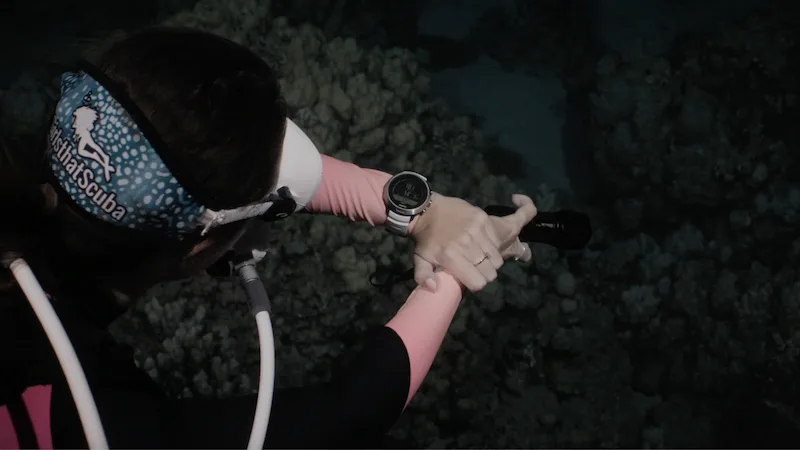 A scuba diver pictured from above in dark water, with a Girls that Scuba blue whale shark print mask strap, white hoses and mask, and a Suunto D5 white dive computer on her wrist.