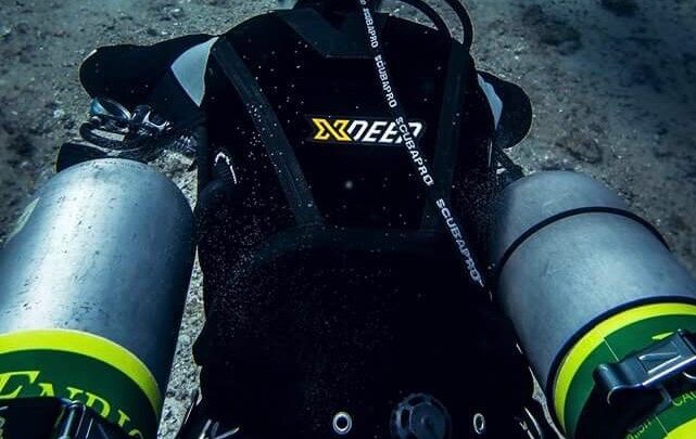 Scuba diver in sidemount configuration with cylinders either side of her. She is swimming away from the camera, and wearing a Girls that Scuba branded mask strap cover.
