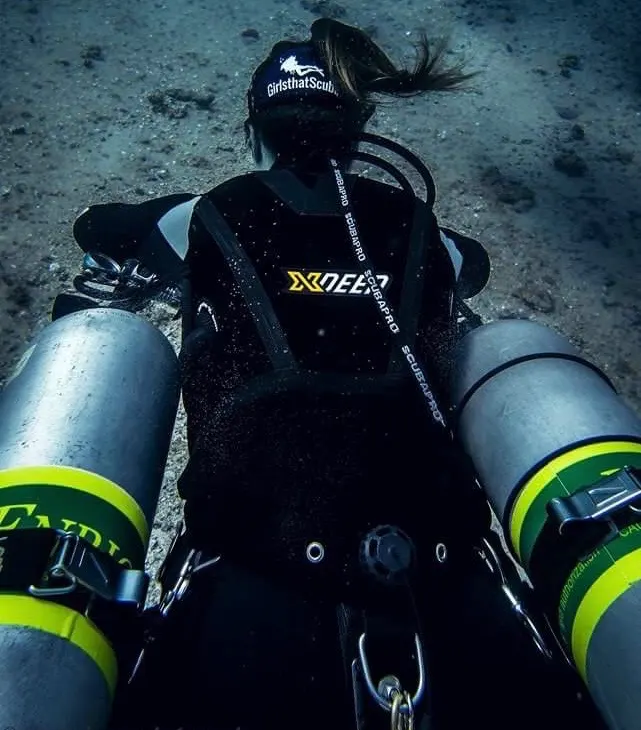 Scuba diver in sidemount configuration with cylinders either side of her. She is swimming away from the camera, and wearing a Girls that Scuba branded mask strap cover. 
