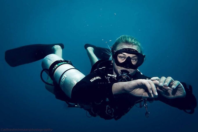 Woman scuba diver looks at the camera in deep blue water. She has two cylinders either side of her body, and her hands are close together in front of her. 