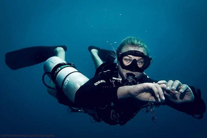 Woman scuba diver looks at the camera in deep blue water. She has two cylinders either side of her body, and her hands are close together in front of her. 