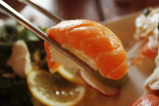 A piece of orange coloured salmon sushi is held up to the camera with metal chopsticks.