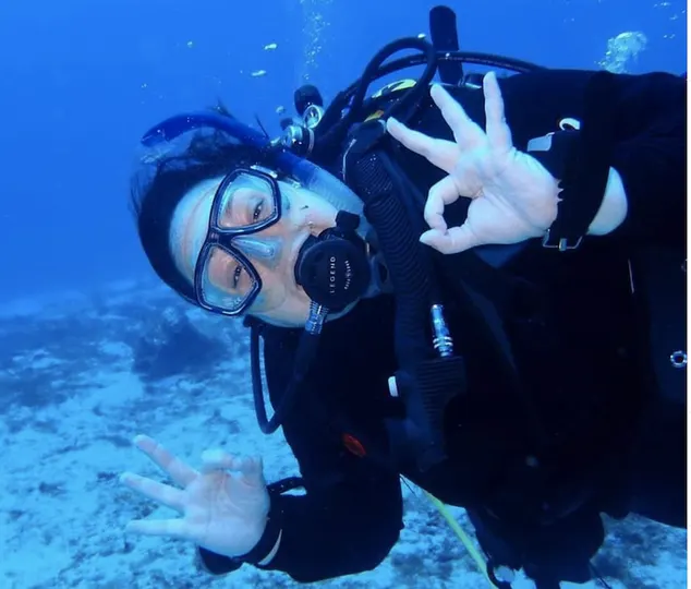 Plus size woman scuba diver is underwater smiling at the camera and holding up an "okay" hand signal 