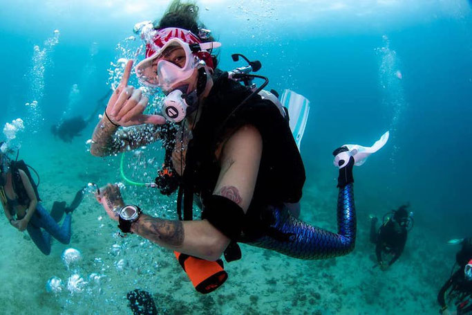 A woman scuba diver looks at the camera and holds up a "hang loose" hand signal, swimming in bright blue water