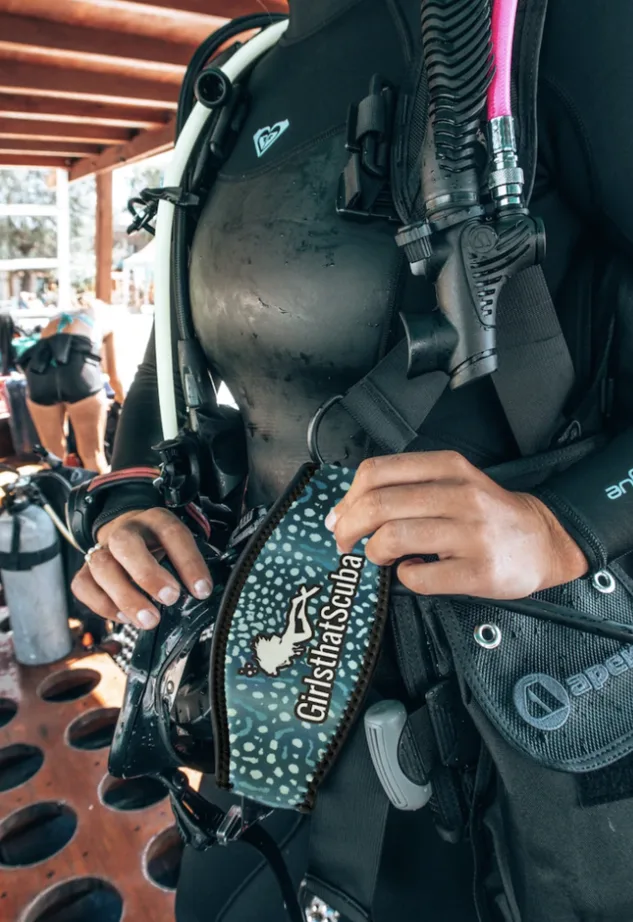 A close up of a female scuba diver wearing black equipment. She is holding a black mask with a Girls that Scuba neoprene mask strap cover with whale shark print.