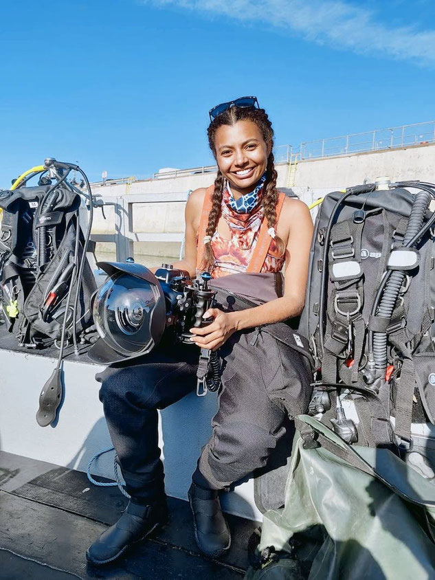 Woman smiles at the camera wearing a Fourth Element drysuit rolled down to the waist and holding a large underwater camera