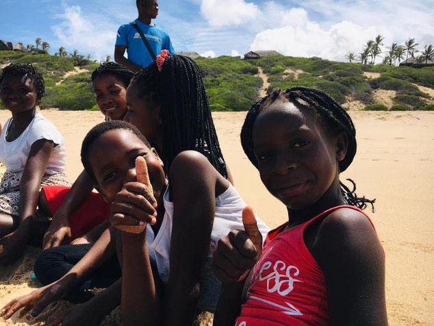 A group of children smile and give a thumbs up at the camera sat on a beach