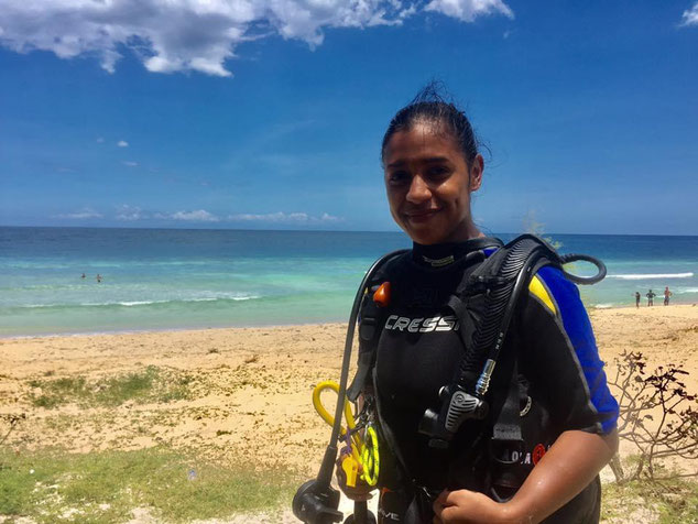 A young woman scuba diver smiles at the camera wearing dive equipment standing on a beach