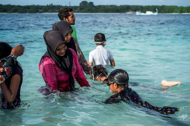 Woman wearing a pink rash vest and black head covering teaches a young girl to swim in shallow ocean
