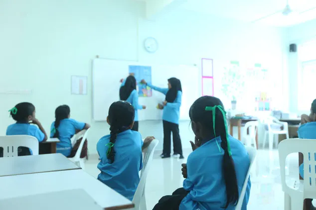 Young girls sit in a classroom, with two older girls presenting at the front of the room