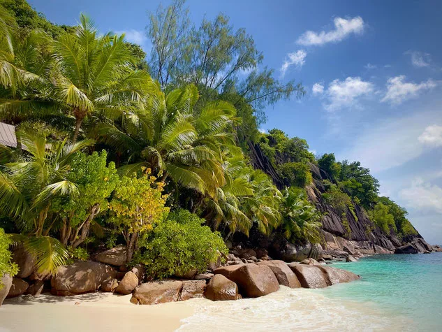 A beach on Mahé Island, bright white sand and turquoise water in foreground, large boulders and lush green vegetation and palm trees in the background