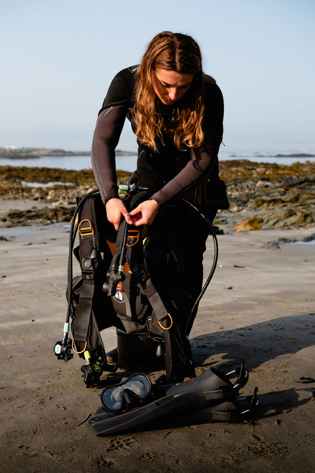 A woman stands on a beach, with scuba equipment in front of her. She is leaning over and adjusting the equipment.
