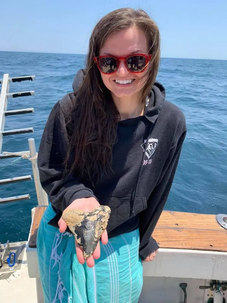 A woman diver holds up a large megalodon tooth fossil to the camera. She is smiling, wearing a black hoodie, red sunglasses, and a blue sarong. She is on a boat with bright blue water behind her.  