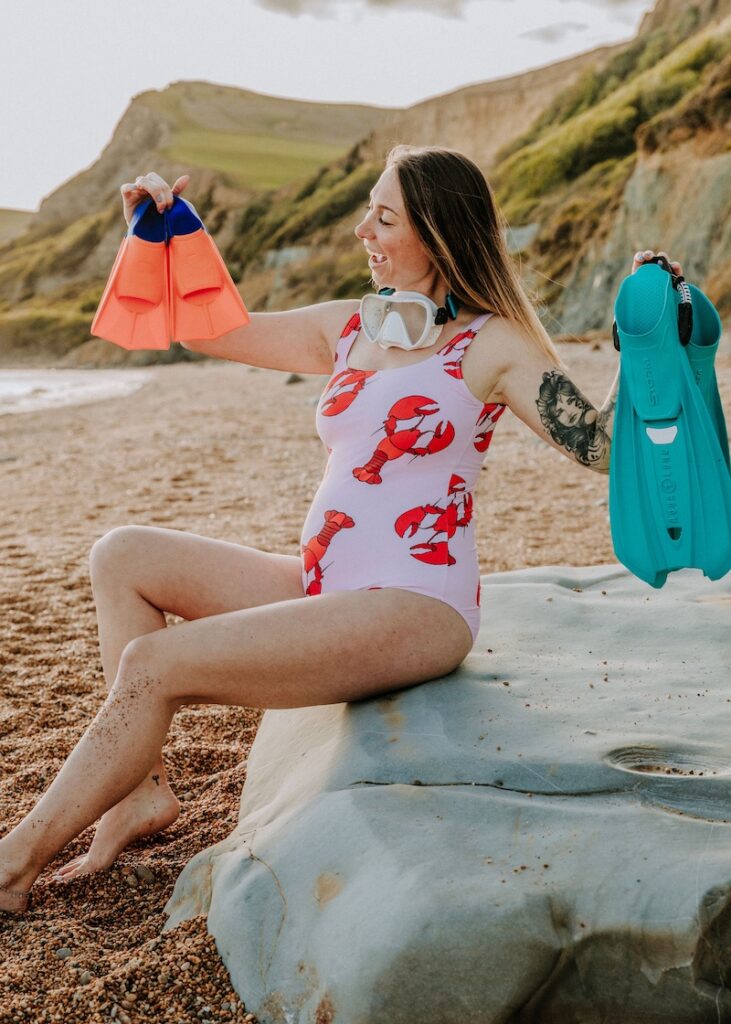 Girls that Scuba founder Sarah sits on a large rock at a beach. She is holding up an adult pair of fins in one hand and a baby pair in the other, smiling at the baby fins. She is wearing a lobster-print swimsuit with a visible baby bump.