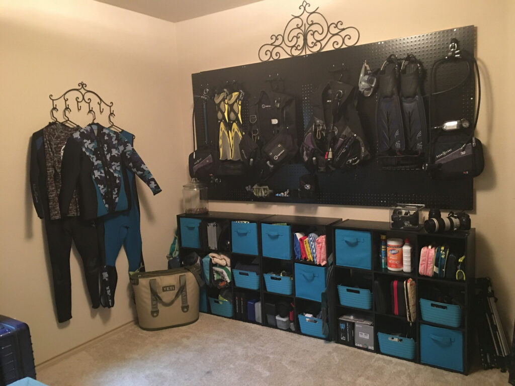 Scuba Gear Storage room, with wetsuits hung on a wall to the left, BCDs and small accessories mounted on a peg board, and other equipment stored in a unit underneath
