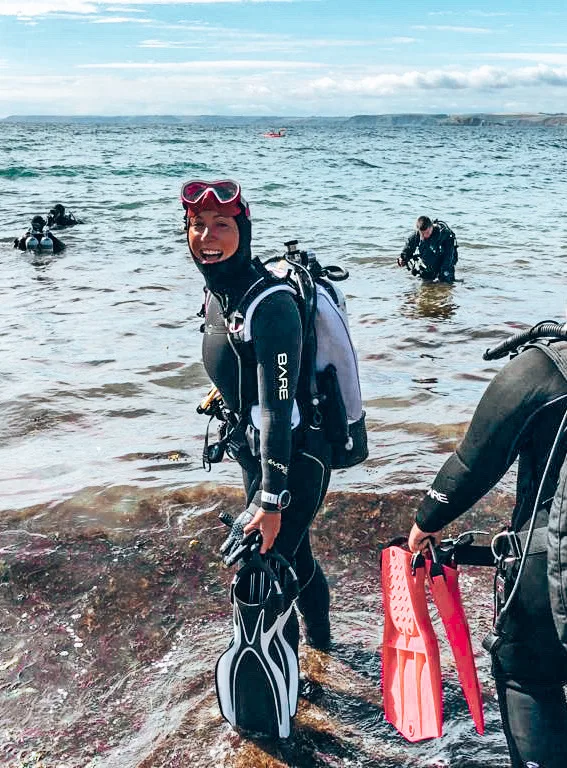 Girls that Scuba founder Sarah goes diving in England. She is smiling at the camera wearing black and white dive equipment with a raspberry pink mask. 