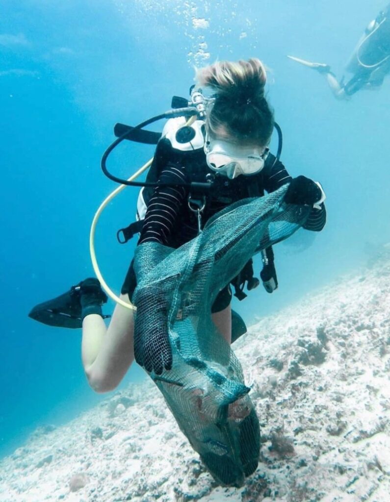 A scuba diver hovers above the ocean floor, holding a mesh bag and collecting underwater litter. 