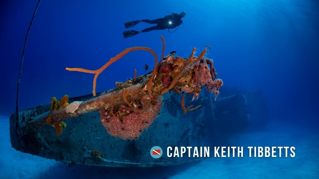 A rusted Soviet shipwreck lays on its side in bright blue water with coral growing on the mast. A scuba diver swims above shining a torch at the camera.