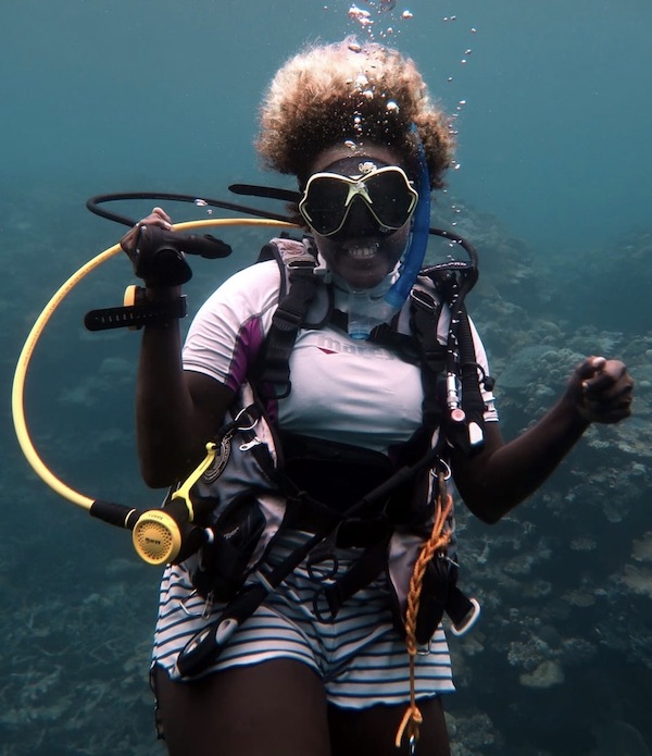 Girls that Scuba Ambassador Euna smiles underwater, holding her regulator out of her mouth. She is wearing a white rash guard and striped shorts, and her tightly curled blonde hair floats around her. 