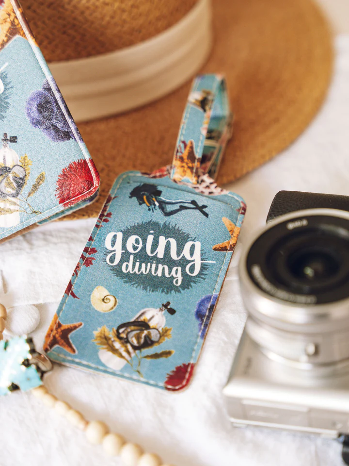 A turquoise luggage tag printed with ocean images including a woman diver, shells, and coral, lays on a table alongside a camera and a sun hat