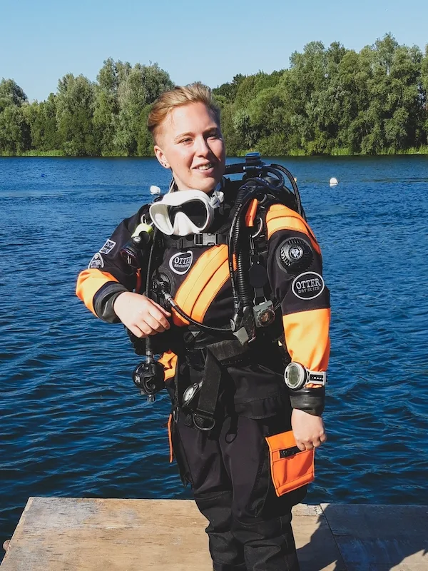 Girls that Scuba Ambassador Grace smiles into the distance, standing on a jetty next to a lake. They are wearing a black and orange drysuit and scuba equipment. 