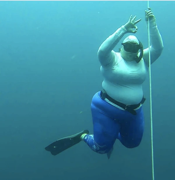 Girls that Scuba Ambassador Irene is freediving holding onto a line, and giving an "OK" hand signal to the camera. She is wearing a long sleeved white rash guard and bright blue leggings.