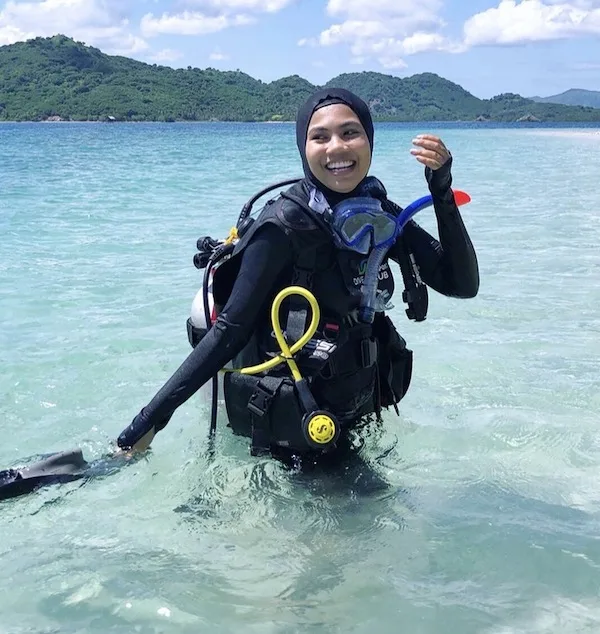 Jessy smiles into the distance. She is stood in waist deep water wearing scuba equipment, and a black rash guard with a head covering.