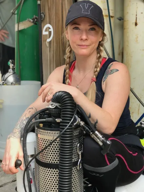 Maria smiles at the camera, her blonde hair is in two braids and she is wearing a black basebell cap. She is leaning over a scuba rebreather.