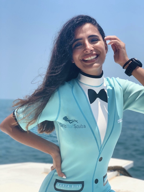 Mariam smiles at the camera, with one hand tucking her long dark hair behind her ear. She is wearing a turquoise wetsuit with a tuxedo design and the Girls that Scuba logo on the front. 