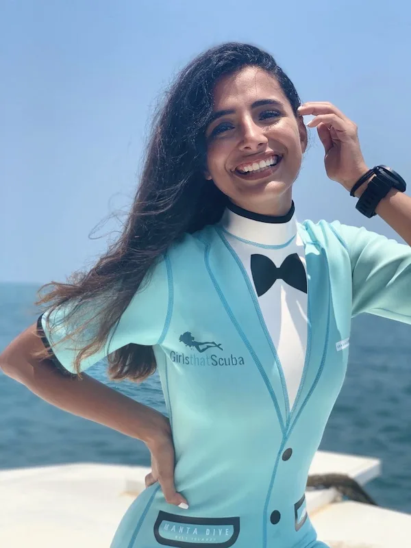 Mariam smiles at the camera, with one hand tucking her long dark hair behind her ear. She is wearing a turquoise wetsuit with a tuxedo design and the Girls that Scuba logo on the front.