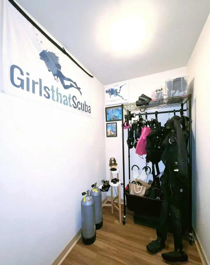 Scuba diving gear storage room with equipment hanging, stored on shelf at the top, and in a box at the bottom. A Girls that Scuba flag is displayed on the wall.