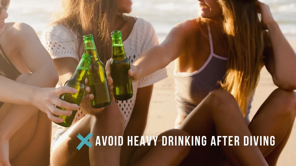 Three women drinking beer on the beach at sunset. Overlaid white text reads "avoid heavy drinking after diving", with a teal cross symbol. 