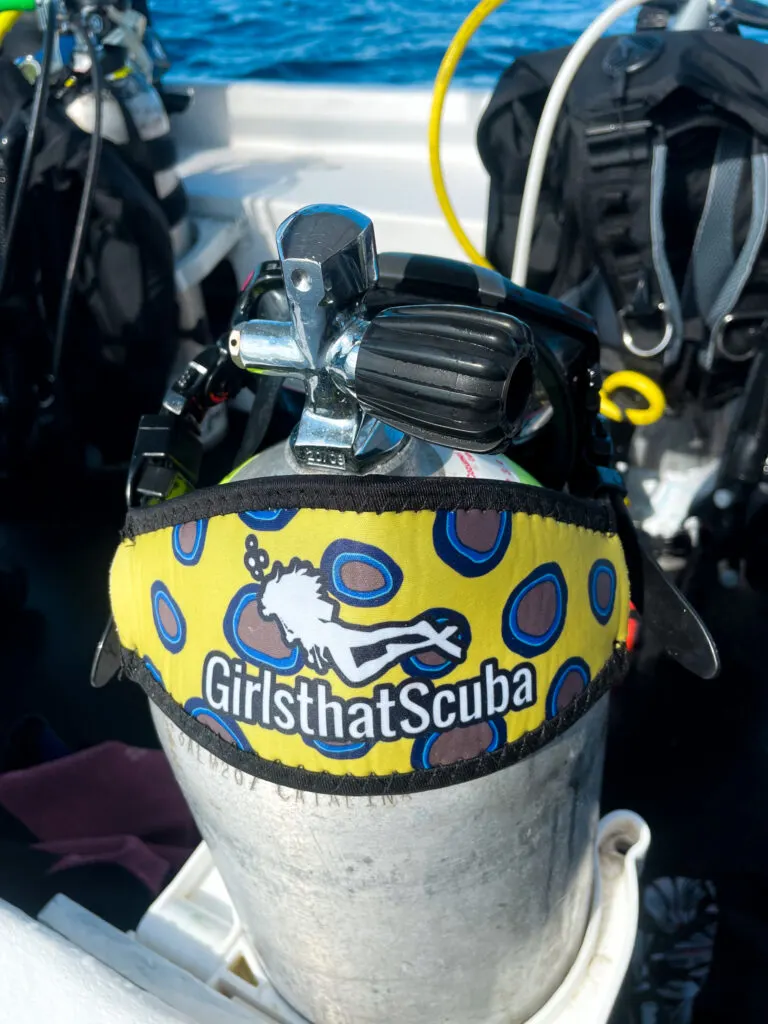 A scuba mask sitting over a dive cylinder, with a neoprene mask strap cover printed with a yellow print with blue rings to imitate an octopus, and the Girls that Scuba logo of a woman diver silhouette.