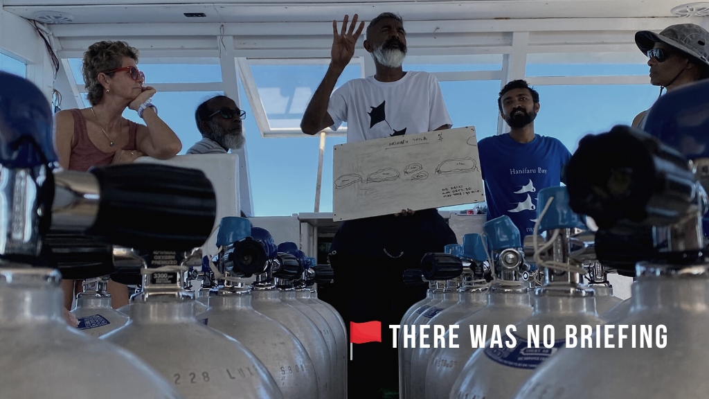 A dive instructor hosts a dive briefing on a boat, he is holding a whiteboard with a map and scuba cylinders are in the foreground. Overlaid white text reads "There was no briefing" with a red flag icon. 