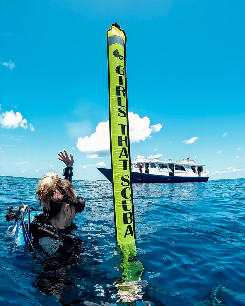 A diver floats at the surface and waves at a dive boat in the background. She is holding up a bright yellow inflatable tube, a DSMB, which signals her position at the surface. The DSMB is labelled with black text reading "GIRLS THAT SCUBA". 