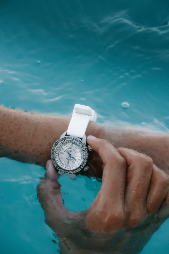 A dive watch with a white strap, silver face, and blue accents, is around a woman's wrist. Her arm is in the surface of blue ocean, and her other hand is adjusting the bezel. 