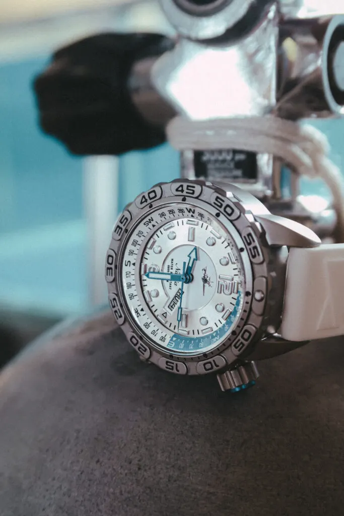 A dive watch with a white strap, silver face, and blue accents, is wrapped around the neck of a scuba diving cylinder. 