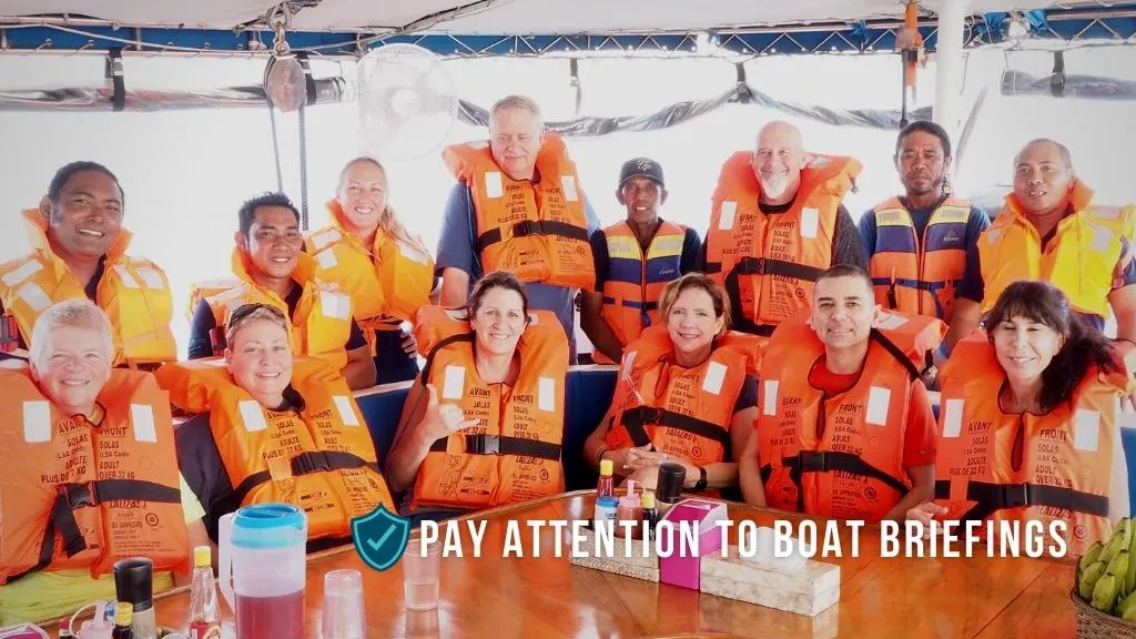 A group of people on a large boat wearing bright orange life jacket buoyancy aids. Overlaid white text reads "pay attention to boat briefings".