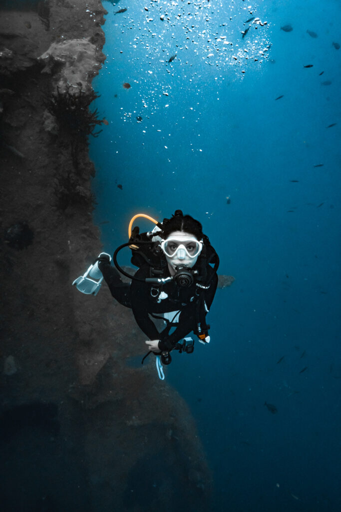 Andi, a scuba diver, looks towards the camera wearing a white mask and fins with black equipment. The SS Coolidge shipwreck found in Vanuatu looms to her left.