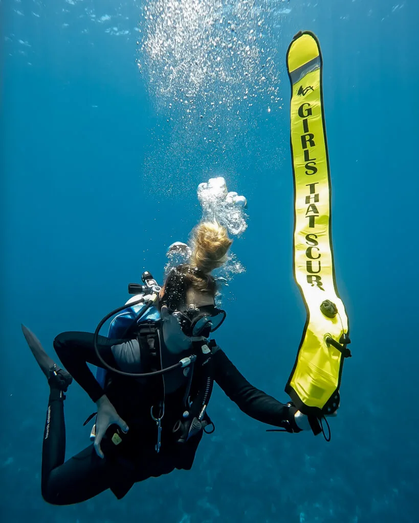 A woman scuba diver is underwater in bright blue water. She is holding a large yellow inflatable tube which reads "GIRLS THAT SCUBA" in black font. 