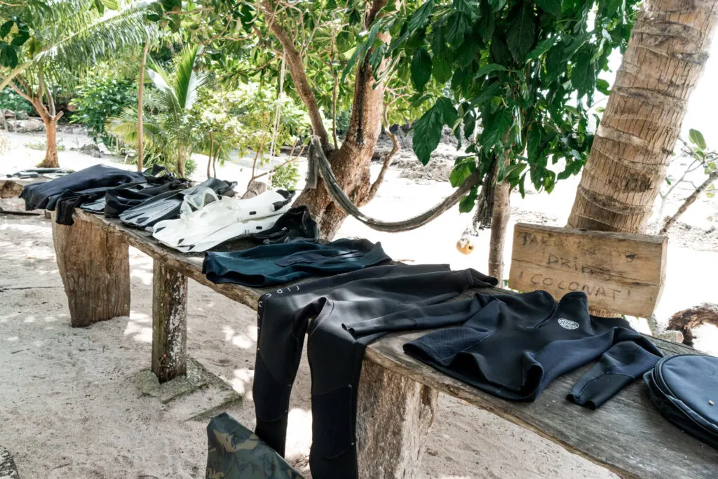 Scuba wetsuits sit on a rustic table surrounded by trees on a sandy white beach. 