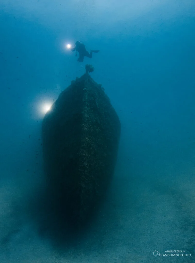 A diver swims above the bow of the Lundy wreck in Gallipoli, Turkey. Another diver's light looms to the left of the large ship. Deep blue water surrounds the shipwreck.