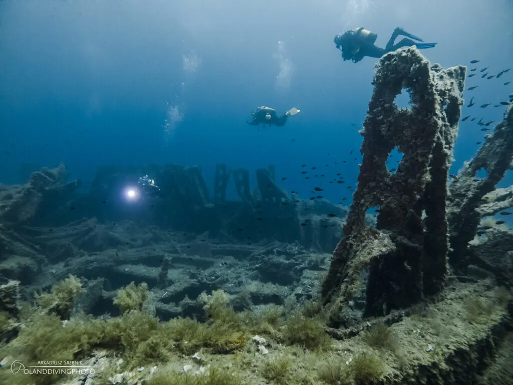 Three people scuba diving in Gallipoli around the HMS Majestic wreck. The wreck is broken up in deep blue water. Algae covers the wreck close to the camera, and fish school to the right of the image.