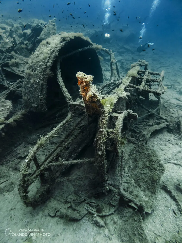 Scuba diving in Gallipoli around the HMS Majestic wreck. The wreck is broken up in deep blue water. A large portion of the ship is fallen in this image, and a school of fish swim above. 