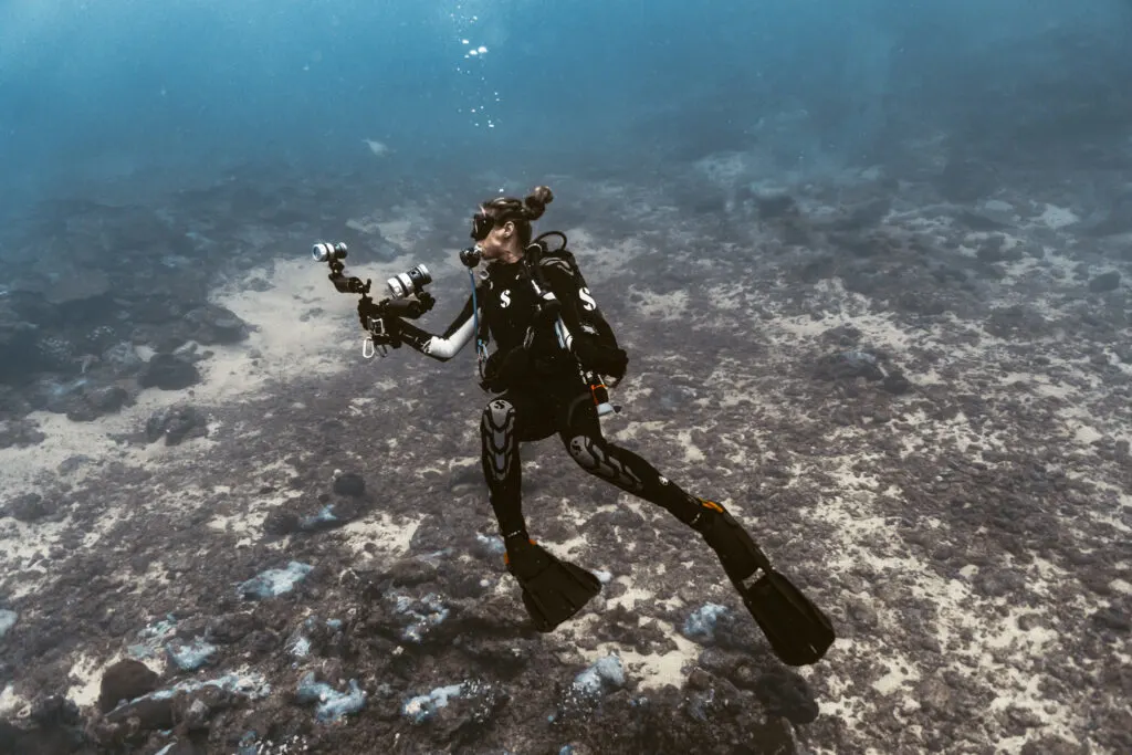 A woman scuba diver swims on her back to the left of the image, across a sandy underwater environment. She carries a large underwater camera. 