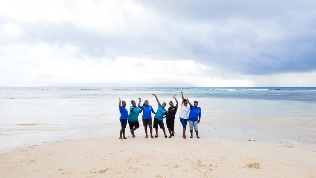 A group of black women stand on a sandy white beach with bright blue ocean in the background. They are smiling and waving at the camera. 