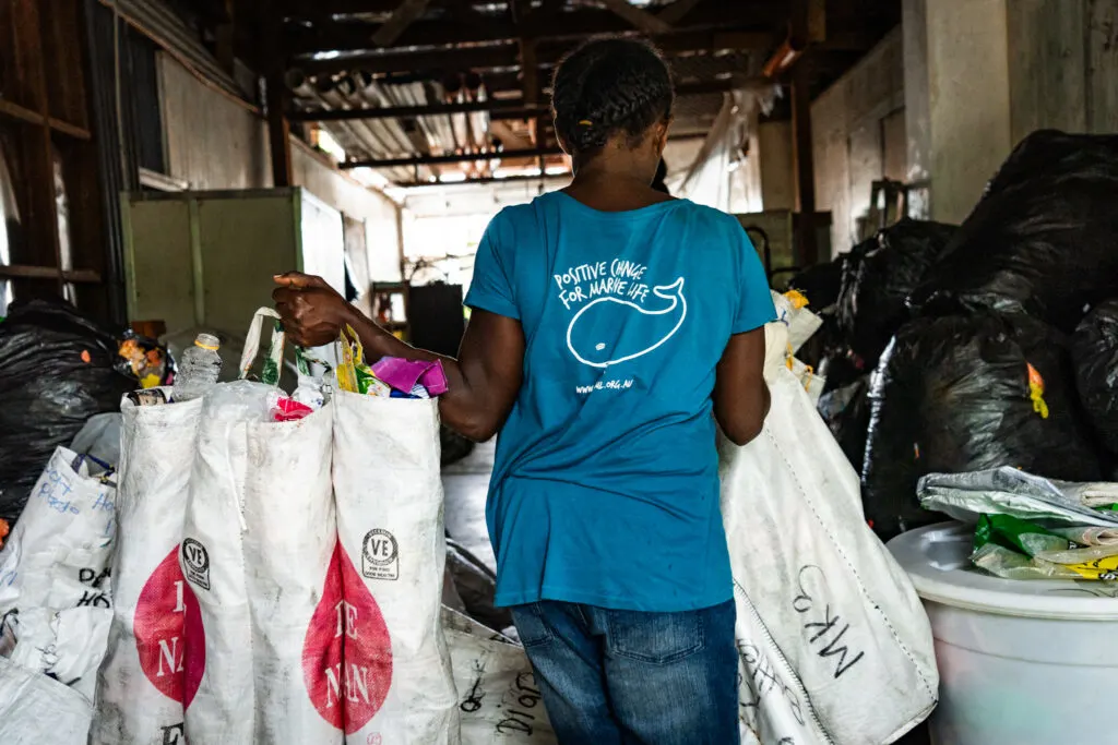 A black woman walks away from the camera, carrying armfuls of rubbish bags sorted for recycling. She is wearing a blue t-shirt which reads "positive change for marine life" with a white outline of a whale. 