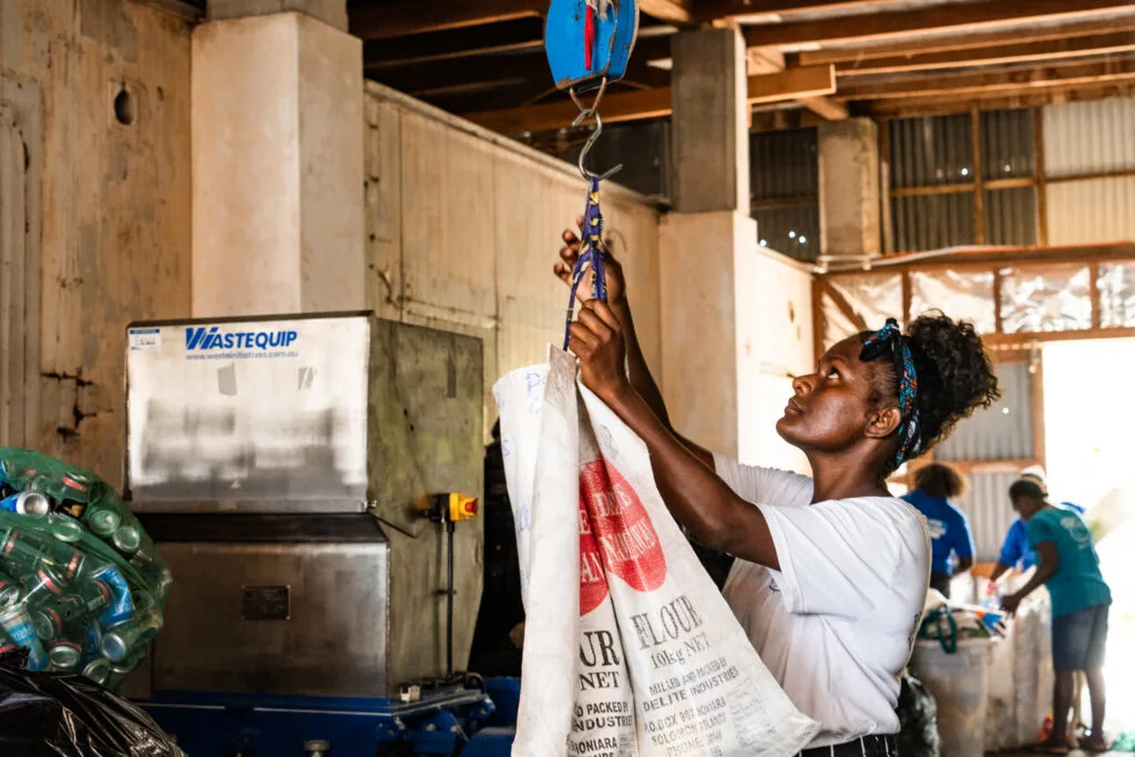 A black woman looks up at a scale as she weighs a large white sack on a hook. She is wearing a white tshirt and a blue scarf in her curly hair. More women sort through rubbish in the background. 