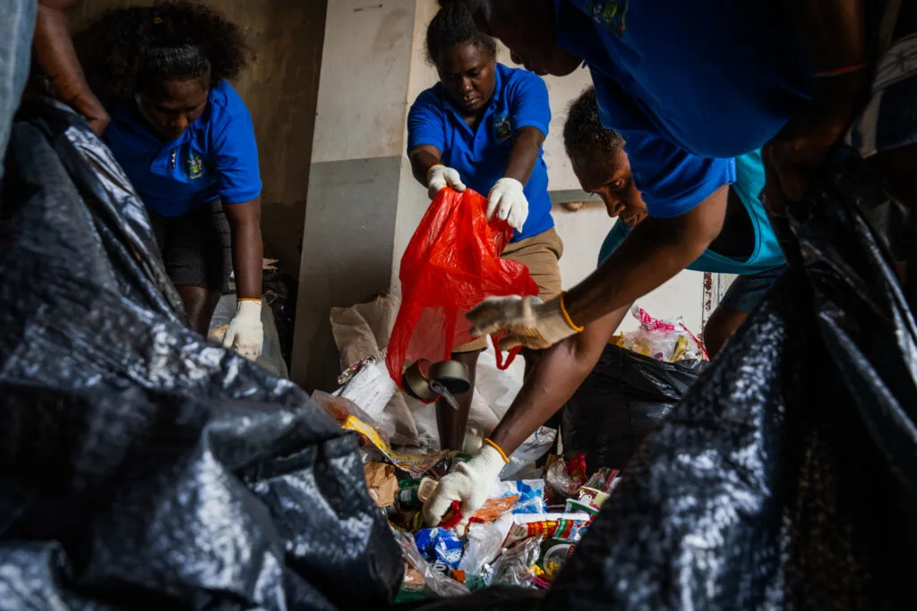A group of black women lean over a pile of trash. They are wearing gloves, pouring more litter in and sorting the pile in the centre.